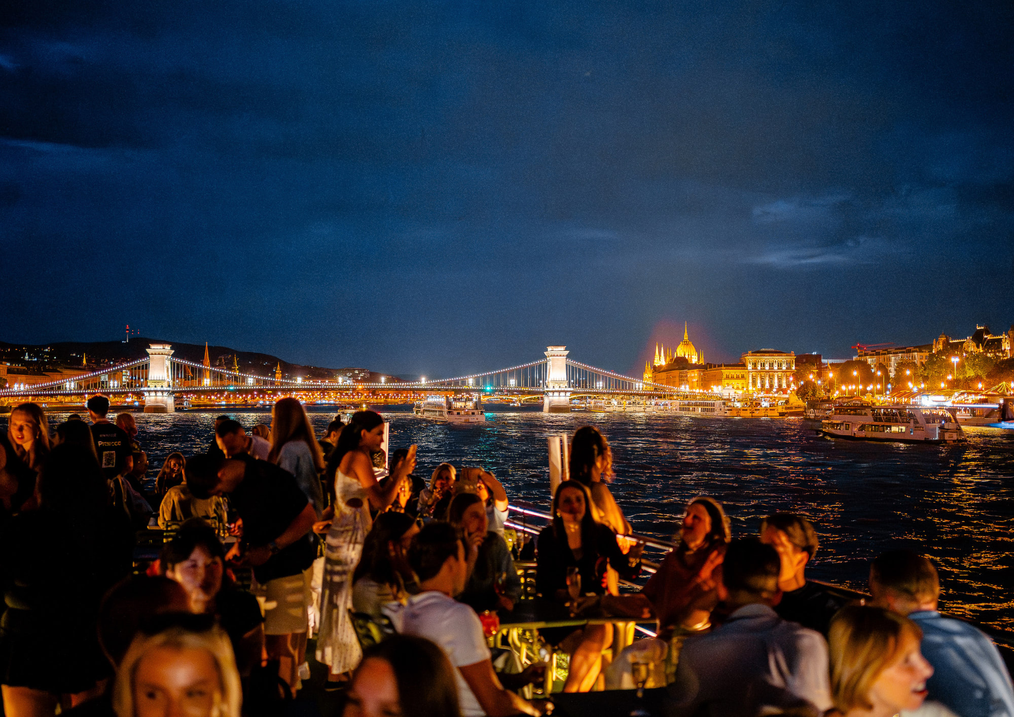 Guests on deck overlooking the illuminated Chain Bridge and Buda riverfront at night.