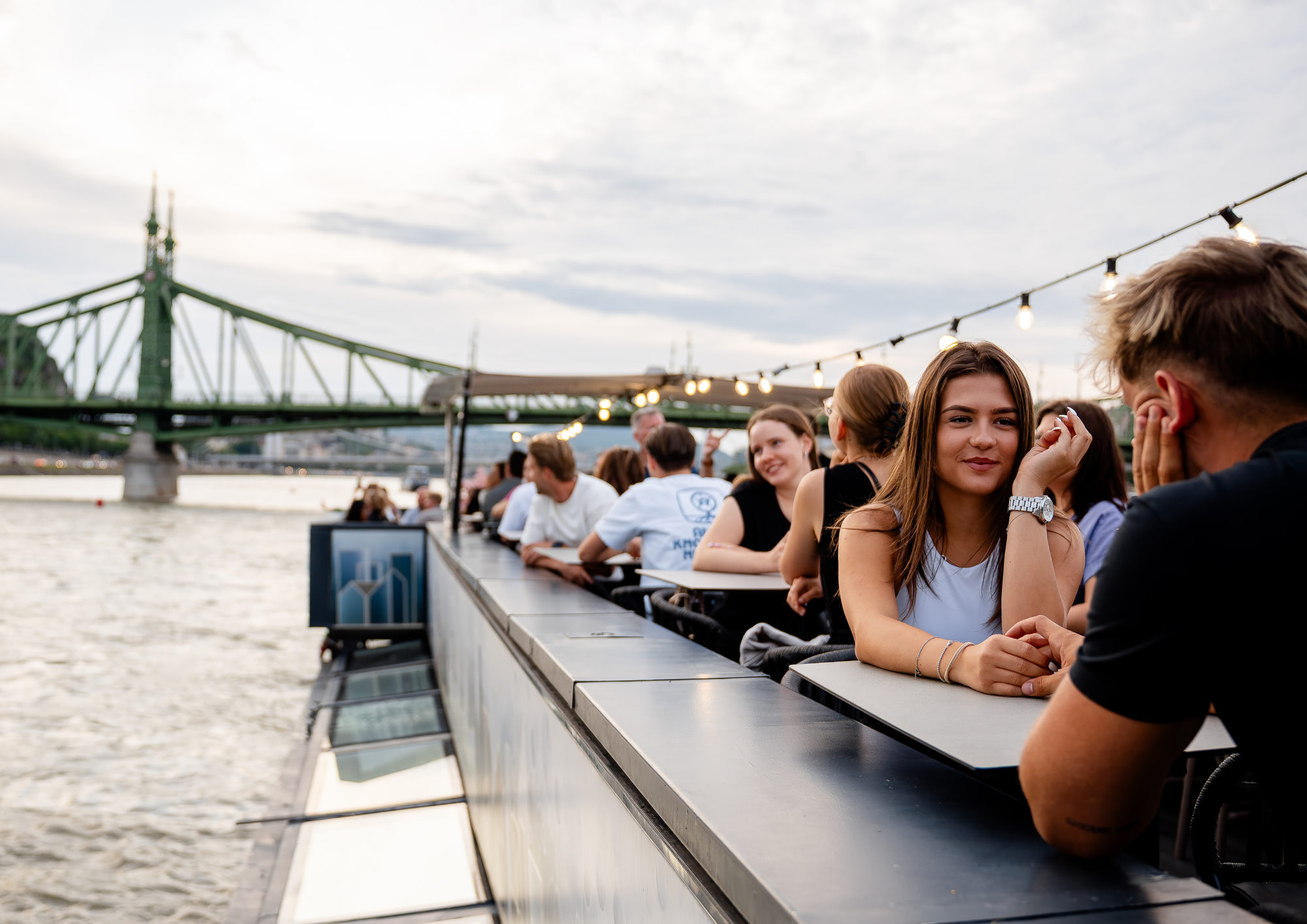 Guests relaxing on the open deck of a Bud By Boat cruise near Liberty Bridge in Budapest.